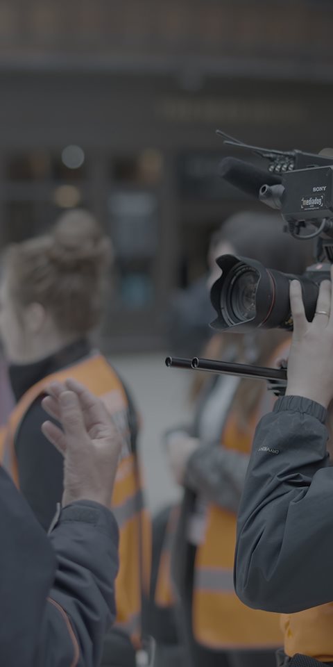 A camera man shooting a video at the train station with multiple people standing and walking around him.